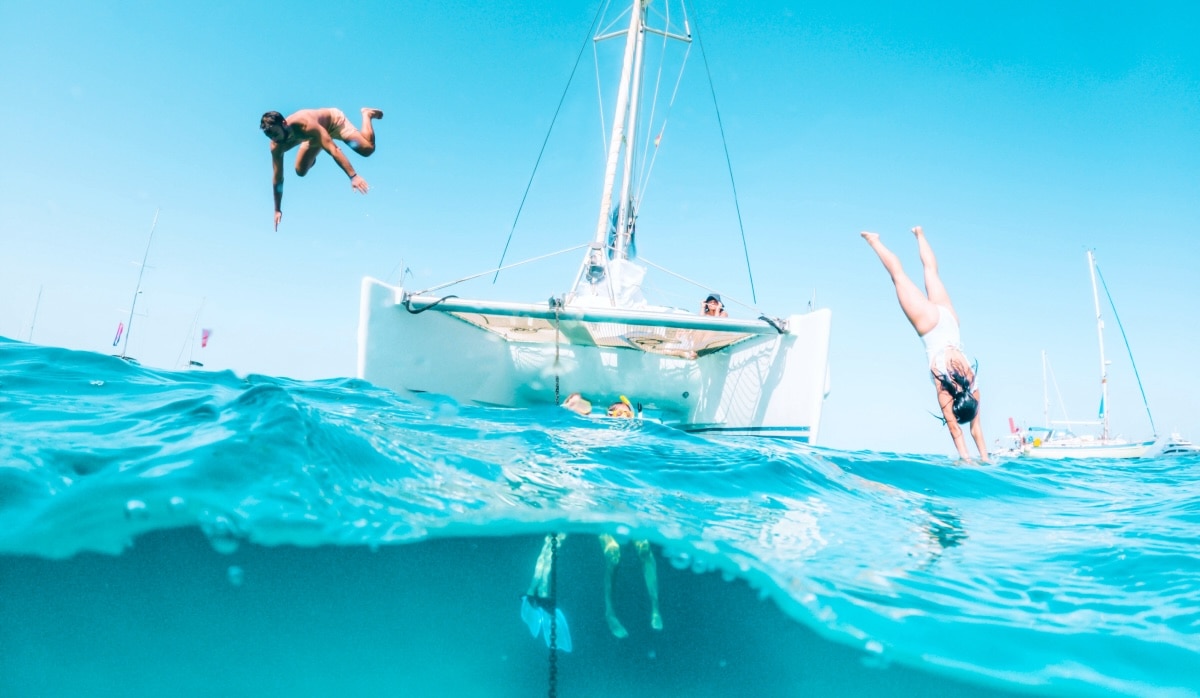 Two people diving off of a sailboat while a friend watches and 2 others are snorkeling nearby.